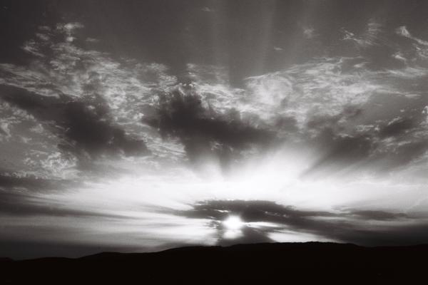 The sun sinks behind the hills, scattering rays through the clouds like an explosion of light and shadow. It’s a simple moment — day becoming night — yet it feels monumental. The contrast is strong, almost cinematic.