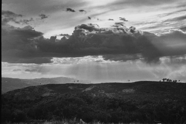 Light breaks through the heavy clouds in scattered beams, touching the hills below with a quiet, almost divine presence. The contrast between the dark sky and the illuminated landscape gives the scene a sense of depth and drama. Nature caught between storm and calm..