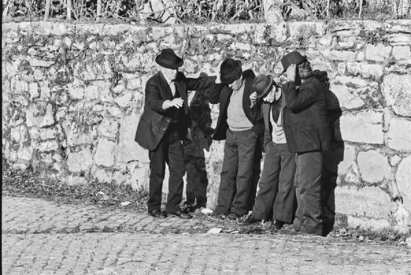A humorous moment frozen in time. Three men instinctively touching their hats while listening attentively to an older gentleman. The sunlight, the cobblestone street, and the stone wall behind them set the perfect stage for this small village scene.